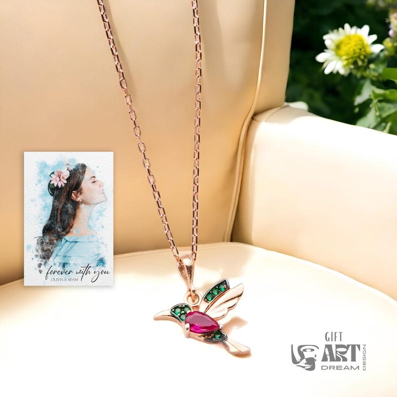 May include: A rose gold necklace with a hummingbird pendant. The pendant features a ruby-colored gemstone and green accents. A small watercolor portrait with the text "forever with you" is visible in the background.