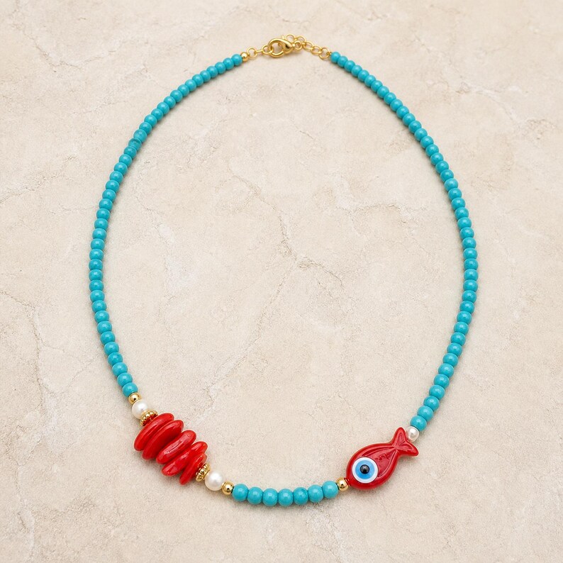 May include: A beaded necklace featuring small turquoise-colored beads, a cluster of four red oval-shaped beads, a single red fish-shaped bead with a blue and white eye design, small white pearls, and gold-toned metal spacers and a clasp.
