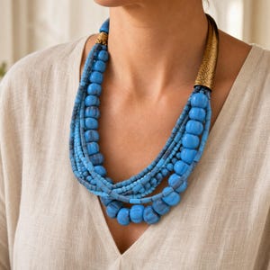 May include: A multi-strand necklace featuring various shades of blue beads in different sizes and textures. The necklace includes a prominent, elongated, etched gold-tone metal cone accent on one side and a smaller, ribbed gold-tone metal bead.