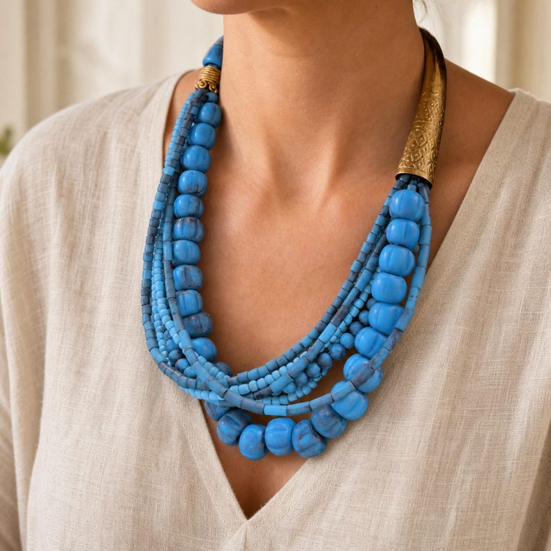 May include: A multi-strand necklace featuring various shades of blue beads in different sizes and textures. The necklace includes a prominent, elongated, etched gold-tone metal cone accent on one side and a smaller, ribbed gold-tone metal bead.