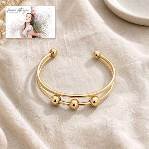 May include: A gold-toned cuff bracelet featuring a double-band design with three polished spherical beads attached to the lower band. In the upper corner, a rectangular card displays a photograph of a couple with the text "forever with you".