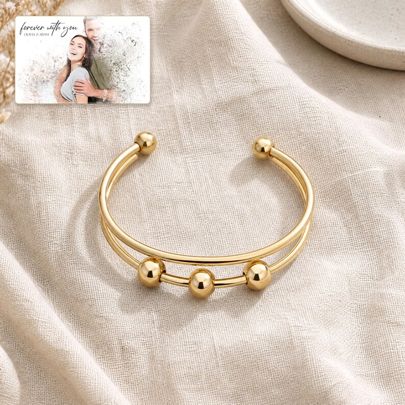 May include: A gold-toned cuff bracelet featuring a double-band design with three polished spherical beads attached to the lower band. In the upper corner, a rectangular card displays a photograph of a couple with the text "forever with you".
