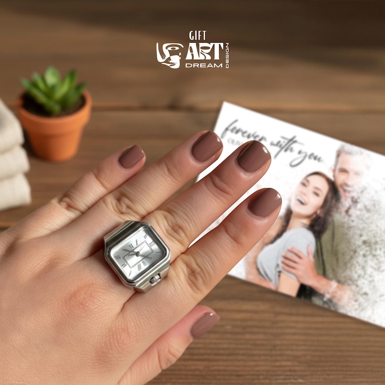 May include: A silver-toned ring watch with a square face is worn on a finger. The watch has a white dial with black hands and Roman numeral hour markers. The image also shows a card with the words "forever with you". The logo "GIFT ART DREAM DESIGN" is also visible.