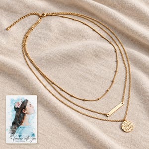 Complete three-layer gold-tone necklace laid on oatmeal linen with its clasp and extension visible. A personalized digital greeting card is shown alongside.