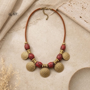 May include: A necklace featuring a brown cord, red porous beads, and gold-toned hammered metal discs. The design includes alternating beads and circular metal pendants arranged symmetrically. A metal clasp is visible at the top of the cord.