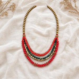 May include: A multi-strand necklace featuring a top section of metallic gold beads that transition into four layered strands. The strands consist of red and dark green faceted beads, small metallic gold spacers, and larger red beads.