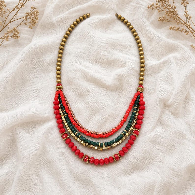 May include: A multi-strand necklace featuring a top section of metallic gold beads that transition into four layered strands. The strands consist of red and dark green faceted beads, small metallic gold spacers, and larger red beads.