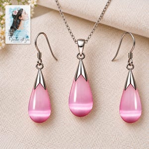 Puede incluir: Un conjunto de joyería que consta de un collar de tono plateado con un colgante en forma de lágrima de piedra ojo de gato rosa y dos pendientes a juego en forma de lágrima con ganchos y tapas metálicas plateadas.