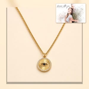 Gold-tone necklace with a round sunburst and black eye detail, light-bordered product view. A personalized greeting-card example appears in the corner.