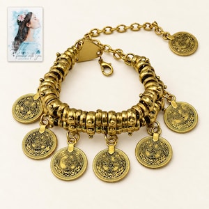 Könnte beinhalten: Ein goldfarbenes Metallarmband mit einem zweireihigen Band aus texturierten, zylindrischen Perlen. Mehrere kreisförmige Münzanhänger mit geprägten Blumen- und Sternmustern hängen am Band. Das Armband enthält eine Verlängerungskette und einen Karabinerverschluss.