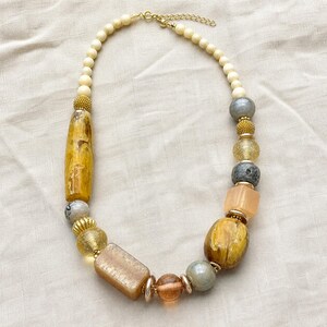 Gift ready presentation of Asymmetrical cream, amber and gray beaded statement necklace with gold spacers in an open jewelry box lined with natural ivory linen