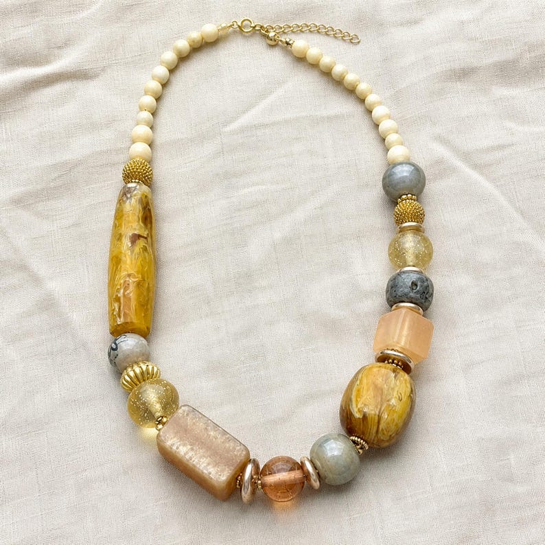 Gift ready presentation of Asymmetrical cream, amber and gray beaded statement necklace with gold spacers in an open jewelry box lined with natural ivory linen