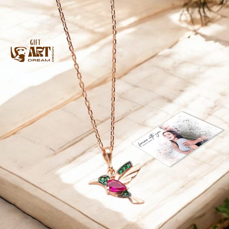May include: Rose gold hummingbird pendant necklace with a ruby-colored gemstone body and emerald green accents on the wings. The necklace hangs on a delicate chain. The image also includes a small card with the text "forever with you".
