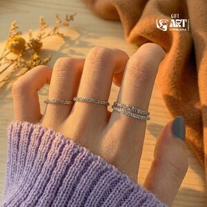 May include: Close-up of a hand wearing three delicate rings. The rings are silver-toned and encrusted with small, sparkling stones. The hand is resting on a purple knitted sweater. The background includes a logo and dried flowers.