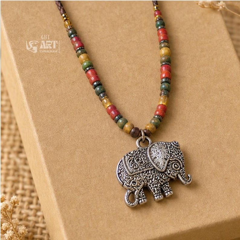 May include: A necklace featuring a silver-toned elephant pendant with intricate embossed patterns, suspended from a cord strung with multicolored beads in shades of red, green, and yellow, with the text GIFT ART DREAM printed in white.