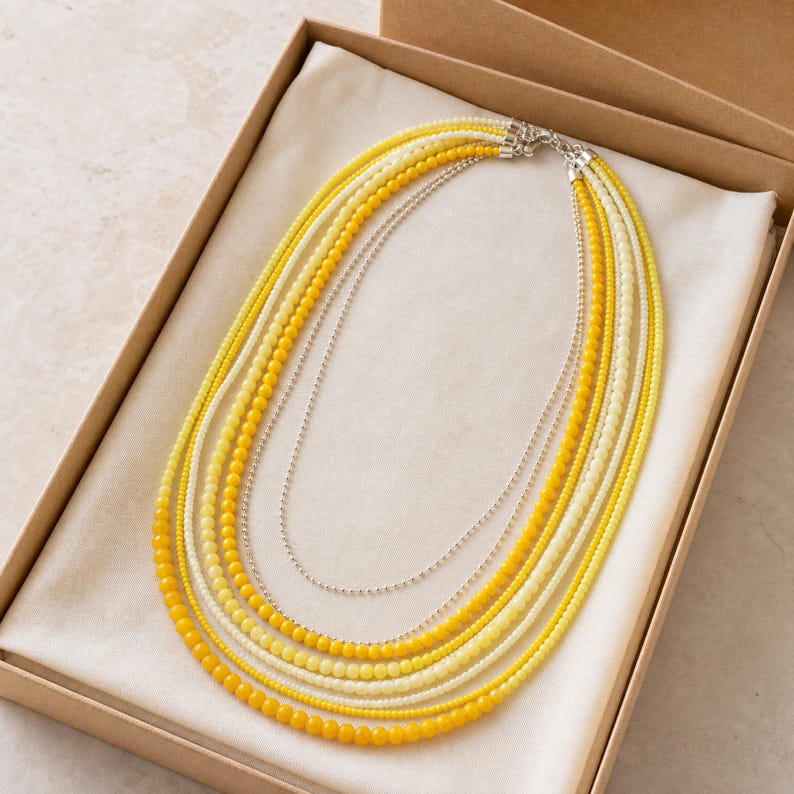 May include: A multi-strand necklace featuring several layers of yellow and cream-colored beads, interspersed with thin silver-toned metallic chains. The necklace is arranged in a circular pattern, showcasing a gradient of warm yellow hues.