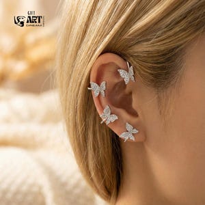 May include: Silver ear cuff set with four butterfly designs, each embellished with small, sparkling crystals. The ear cuffs wrap around the ear. The image also includes the text "GIFT ART DREAM DESIGN".