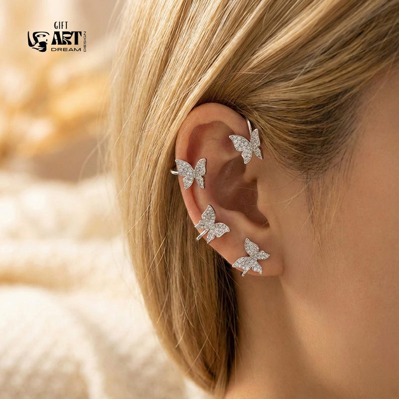 May include: Silver ear cuff set with four butterfly designs, each embellished with small, sparkling crystals. The ear cuffs wrap around the ear. The image also includes the text "GIFT ART DREAM DESIGN".