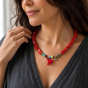 Op de afbeelding: Een ketting met een reeks rechthoekige rode kralen, afgewisseld met kleine goudkleurige tussenstukjes. Het midden bevat een kleine blauwe, gele en witte visbedel, een rood kwastje en extra veelkleurige decoratieve kralen.