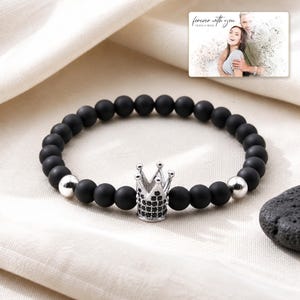 Puede incluir: Una pulsera de cuentas esf&eacute;ricas negras mate, dos cuentas plateadas pulidas y un dije central de corona plateada incrustado con peque&ntilde;as piedras negras. Una tarjeta rectangular muestra una foto de una pareja con el texto forever with you.