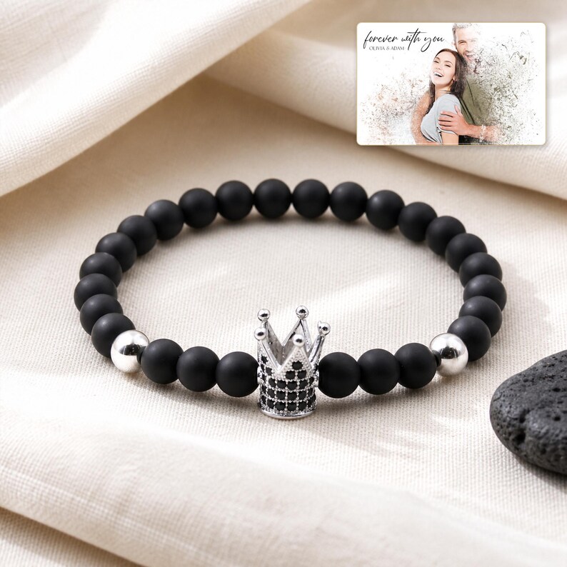 Puede incluir: Una pulsera de cuentas esf&eacute;ricas negras mate, dos cuentas plateadas pulidas y un dije central de corona plateada incrustado con peque&ntilde;as piedras negras. Una tarjeta rectangular muestra una foto de una pareja con el texto forever with you.