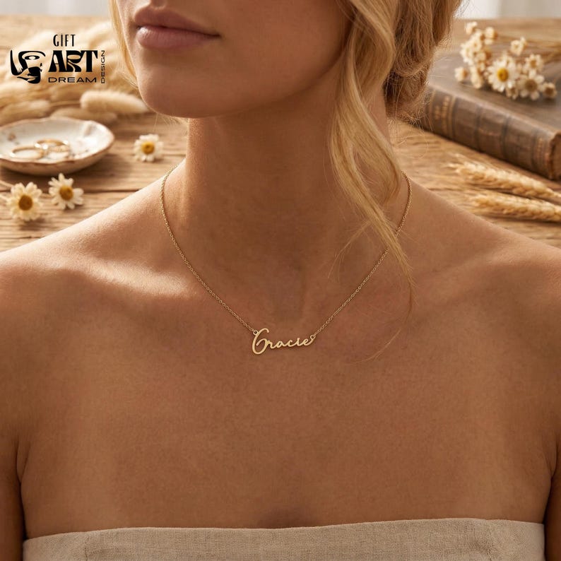 May include: A delicate gold necklace with the name "Gracie" in cursive script. The necklace is displayed on a person's chest, with a wooden surface in the background. The necklace is a piece of jewelry.