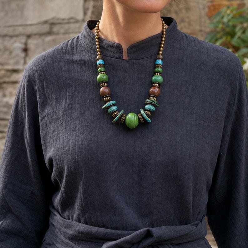 May include: A beaded necklace featuring a central ribbed green bead, flanked by smaller turquoise, brown, and green beads, interspersed with small metallic spacers. The necklace is worn over a dark charcoal gray long-sleeved garment with a high neckline.