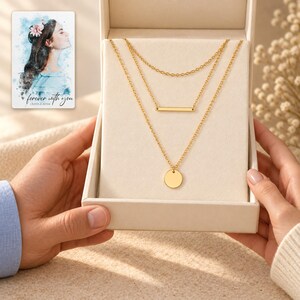 Two adults exchanging an open ivory box containing the complete gold-tone necklace trio; hands stay outside the jewelry. A personalized digital greeting card is shown alongside.