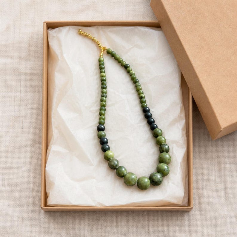 May include: A beaded necklace featuring a gradient of round green and dark charcoal beads, arranged in a graduated size pattern from smaller beads at the top to larger beads at the bottom, finished with a gold-toned metal clasp and chain.