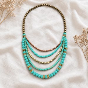 May include: A multi-strand necklace featuring a top row of small metallic bronze beads. Below are four cascading strands composed of turquoise-colored round beads, small metallic bronze cube beads, and gold-toned metal bead caps at intervals.