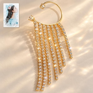 Puede incluir: Un pendiente tipo ear cuff dorado con marco curvo, dos tachuelas de cristal transparente y ocho flecos en cascada de cristales transparentes. Incluye una tarjeta rectangular con una ilustración en acuarela y el texto "forever with you".