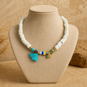 Puede incluir: Un collar con cuentas blancas en forma de cubo, una borla turquesa, una peque&ntilde;a cuenta texturizada de color marr&oacute;n oscuro, una cuenta a rayas de colores con forma de pez y un peque&ntilde;o dije verde y naranja, con espaciadores dorados.