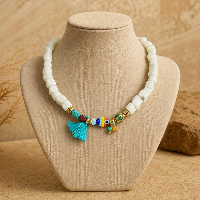 Puede incluir: Un collar con cuentas blancas en forma de cubo, una borla turquesa, una peque&ntilde;a cuenta texturizada de color marr&oacute;n oscuro, una cuenta a rayas de colores con forma de pez y un peque&ntilde;o dije verde y naranja, con espaciadores dorados.