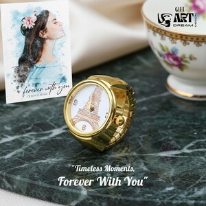 May include: A gold ring watch with a white face and a brown Eiffel Tower design. The watch sits on a dark green marble surface, beside a card with a watercolour portrait and the text "forever with you". The text "Timeless Moments, Forever With You" is also visible.