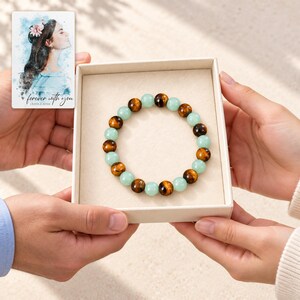 Forest Amber Bead Bracelet shown during a believable gift-giving moment between adults with the jewelry visible. A personalized digital greeting card is shown alongside.