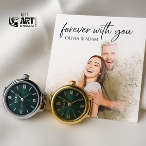 Peut inclure: Deux bagues de montre avec des cadrans verts, une argent&eacute;e et une dor&eacute;e, &agrave; c&ocirc;t&eacute; d'une carte avec le texte "forever with you" et une photo d'un couple. Les bagues de montre ont des chiffres romains et la marque "HAIDI".