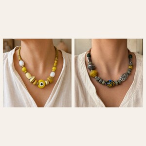May include: Two necklaces featuring varied beads. The left necklace has yellow, white, and gold-toned beads with a central yellow evil eye charm. The right necklace displays grey, yellow, and textured silver-toned beads with a central patterned bead.