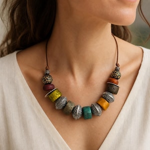 May include: A necklace featuring a brown cord strung with a variety of beads, including textured silver-toned metal beads, smooth round beads in shades of yellow, green, turquoise, and brown, and black beads with a gold-coloured leopard print pattern.