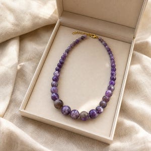 May include: A beaded necklace featuring round, polished purple amethyst stones that graduate in size from smaller beads near the gold-toned metal clasp to larger beads at the centre, arranged in a circular shape within a light-coloured box.