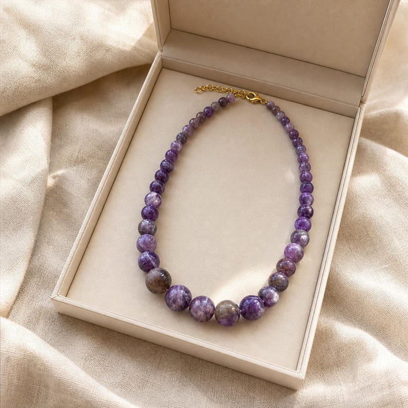 May include: A beaded necklace featuring round, polished purple amethyst stones that graduate in size from smaller beads near the gold-toned metal clasp to larger beads at the centre, arranged in a circular shape within a light-coloured box.