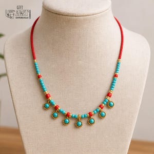 May include: A necklace featuring a red cord base accented with alternating turquoise and red beads, interspersed with gold-toned metal spacers. The lower section includes six dangling circular charms with turquoise centers and gold-toned beaded edges.