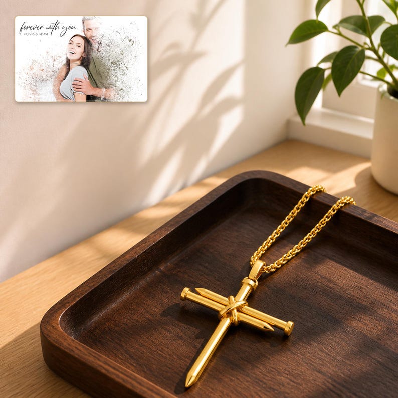 K&ouml;nnte beinhalten: Eine goldfarbene Kreuz-Halskette mit einem Anh&auml;nger aus zwei gekreuzten, miteinander verbundenen N&auml;geln an einer passenden goldfarbenen Kette. Im Hintergrund ist eine rechteckige Karte mit dem Text "forever with you" und "OLIVIA & ADAM" zu sehen.