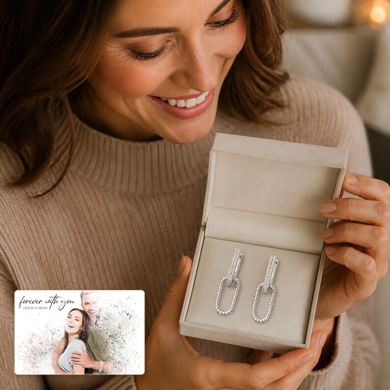 May include: A pair of silver-toned drop earrings featuring an elongated oval link design with a textured, beaded surface. The earrings are presented inside a light beige, open jewellery box. A small rectangular card with the text "forever with you" is visible.