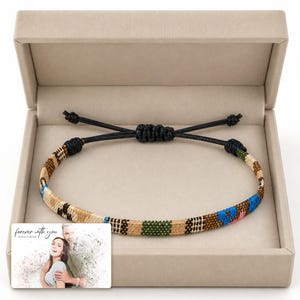 May include: A woven bracelet featuring a multi-coloured geometric pattern in shades of tan, brown, green, and blue, with a black adjustable cord closure. The bracelet is displayed in a beige box next to a card with the text "forever with you."