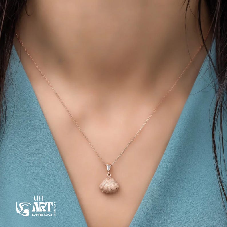 Op de afbeelding: Een delicate ros&eacute;gouden ketting met een schelpenhanger. De hanger is gedetailleerd en de ketting is fijn. De ketting wordt getoond tegen een blauwe achtergrond. De tekst "GIFT ART DREAM DESIGN" is ook zichtbaar.
