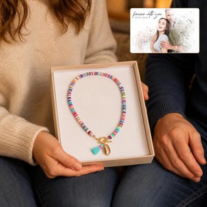 May include: A necklace made of multicolored flat circular beads featuring a gold-toned toggle clasp, a small light blue tassel, and a gold-toned seashell charm, presented in a white gift box with a card showing a couple and the text forever with you.