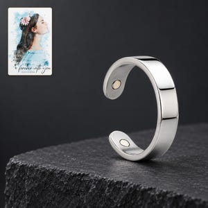 Reflective silver-tone open ring on a charcoal stone style plinth under soft studio lighting. A personalized digital greeting card is shown alongside.