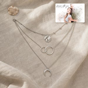 May include: A silver-toned layered necklace featuring three distinct chains. The top chain has a small globe pendant, the middle chain displays two interlocking circles, and the bottom chain holds a crescent moon charm. A photo card reads "forever with you".