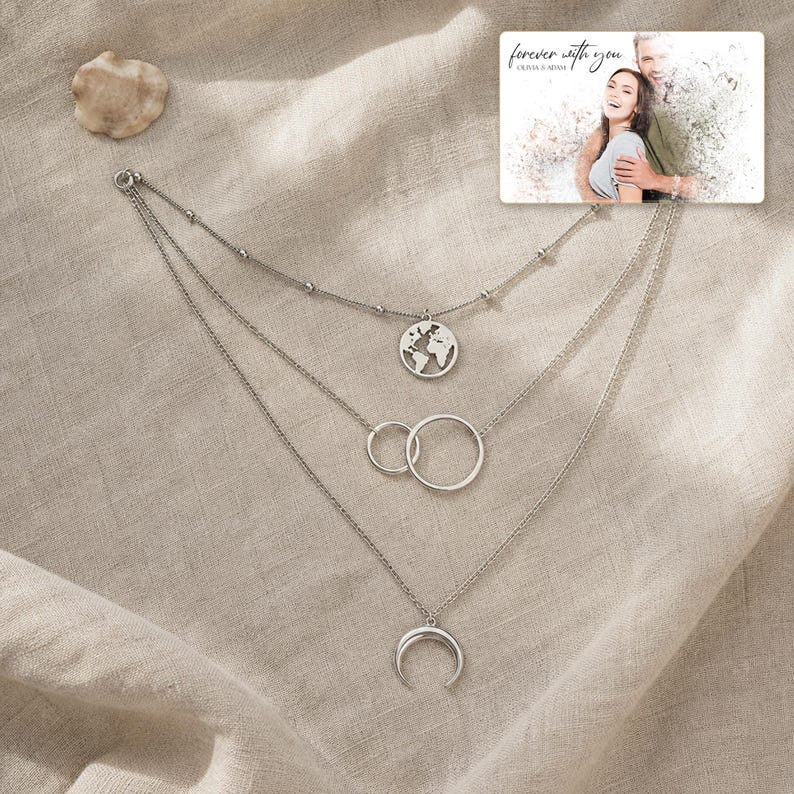 May include: A silver-toned layered necklace featuring three distinct chains. The top chain has a small globe pendant, the middle chain displays two interlocking circles, and the bottom chain holds a crescent moon charm. A photo card reads "forever with you".