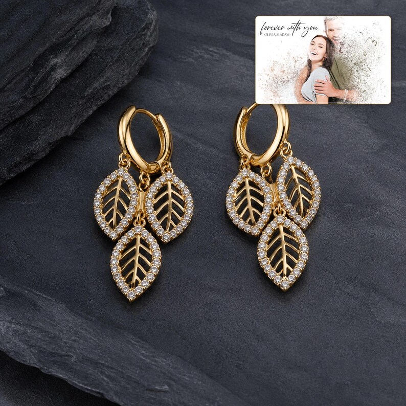 May include: A pair of gold hoop earrings, each featuring three dangling leaf-shaped charms encrusted with small clear stones. An inset photo shows a couple with the text "forever with you OLIVIA & ADAM" above them, set against a dark gray background.